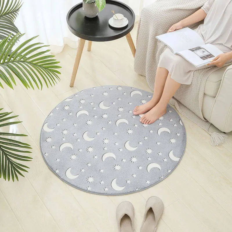 Round grey rug with moon and star patterns on a light wood floor, with a person sitting on a chair next to it.