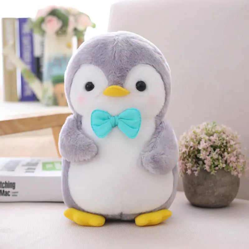 Plush penguin toy with a blue bow tie on a surface with books and plants in the background.
