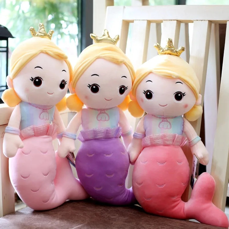 Three plush mermaid toys with blonde hair and crowns on a wooden surface.