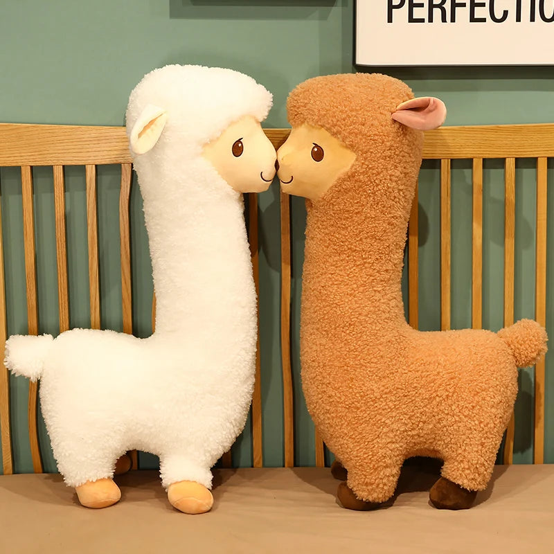 Two plush alpaca toys, one white and one brown, sitting on a bed.
