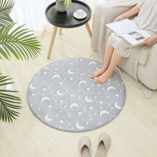 Round grey rug with moon and star patterns on a light wood floor, with a person sitting on a chair next to it.