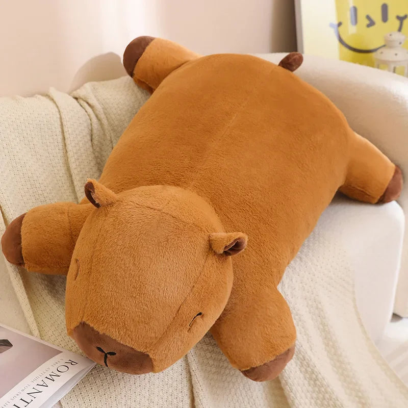 Large brown capybara plush toy lying flat on a sofa, designed as a soft floor and lounge cushion.