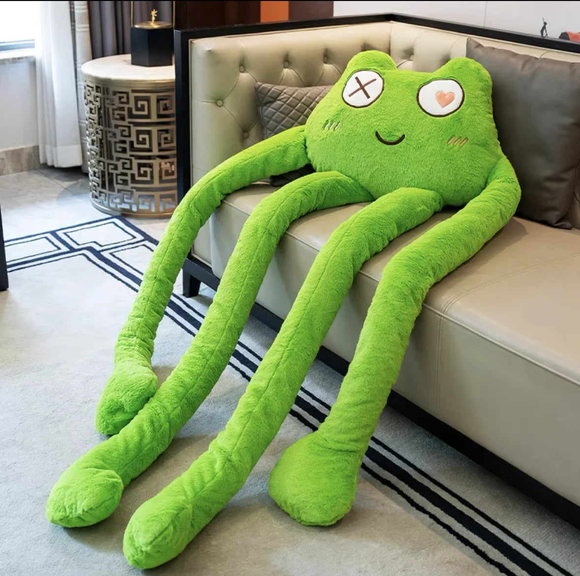 Green long-legged frog plush toy draped across a sofa with playful stitched details