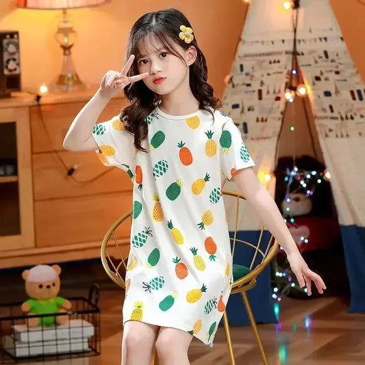 Young girl wearing a pineapple-patterned nightie in a room with a wooden cabinet and decorative lights.